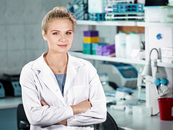 A woman in a white coat with her arms crossed, in front of shelves of laboratory equipment
