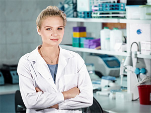 A woman in a white coat with her arms crossed, in front of shelves of laboratory equipment