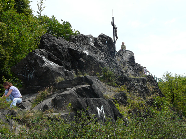 A rock outcrop with a cross embedded in it. Tourists are resting on the rocks. Around it are bushes and trees