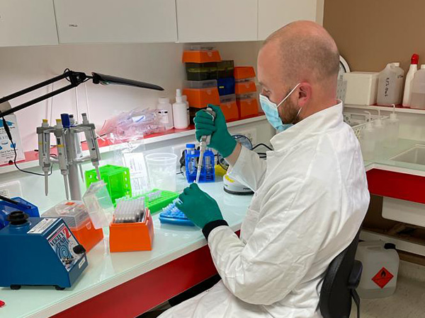 A man in a white coat sits at a lab table. He uses a pipette to inject liquid into a small test tube