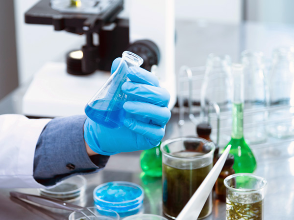 Blat zastawiony sprzętem laboratoryjnym. Na pierwszym planie dłoń w lateksowej rękawiczce trzyma kolbę z niebieskim płynem