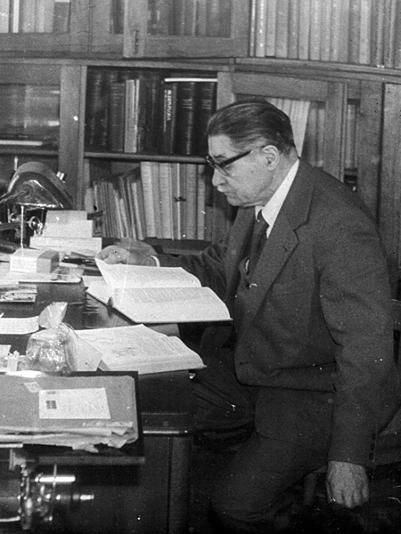 A black and white photo of an elderly man with glasses reading a thick book at his desk. Bookshelves in the background