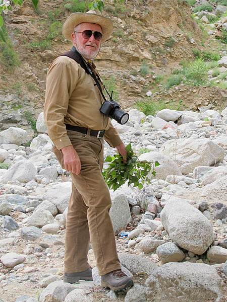 A man in a hat, sunglasses, a camera around his neck and a plant in his hand stands among rocks and sparse vegetation