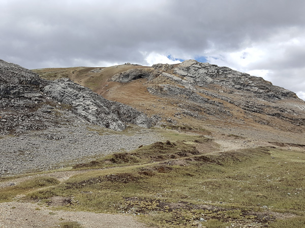A mountain with gentle slopes partially covered with sparse grass vegetation. In the foreground a winding rocky road