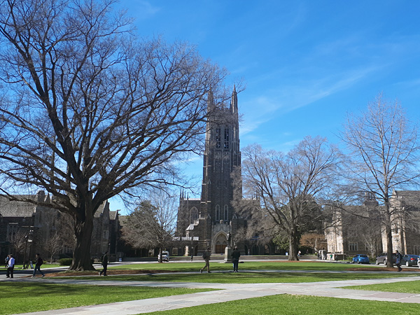 University buildings with a neo-Gothic chapel towering over them. In the foreground a park with a low-cut lawn