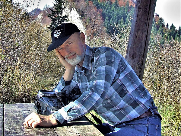 A man with a white beard and a checked shirt sits under a wooden tourist shelter with his head resting on his hand