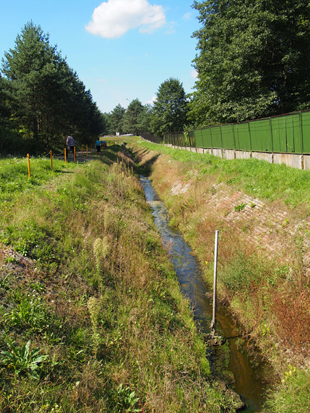 A narrow watercourse with banks lined with openwork concrete slabs. A metal fence runs along one of the banks