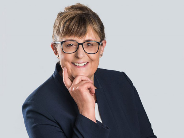 Kobieta w ciemnej garsonce, okularach i upiętych z tyłu włosach. Podpiera ręką głowę i uśmiecha się w kierunku obiektywu