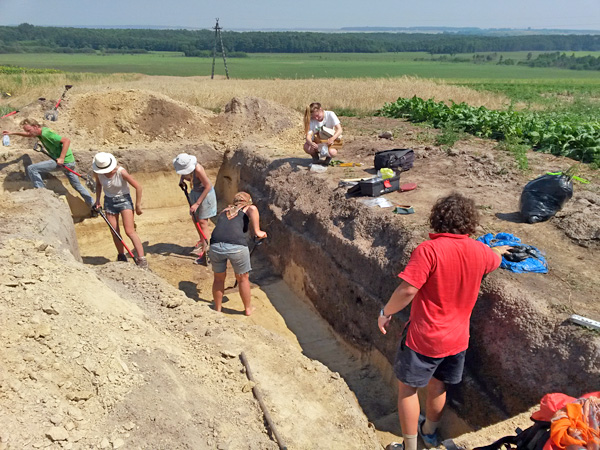 Several people working on deepening a rectangular hole with an agricultural and forest landscape in the background
