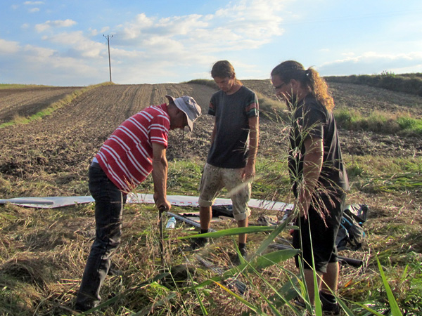 Three men in front of a ploughed field. One of them drives a metal auger into the soil and two others watch