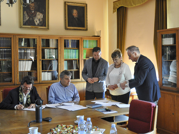 Two men sign documents spread out on a wooden table. Next to them, three other people wait their turn