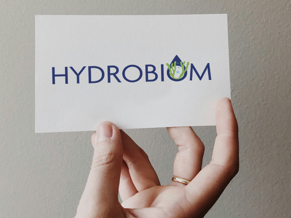 Woman's hand holds a card with a logo containing the navy blue inscription Hydrobiom and a drawing of algae
