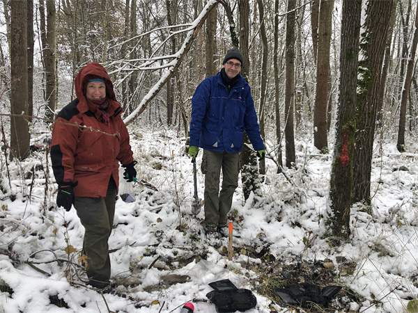 A woman and a man stand in a snowy forest in winter jackets. At their feet are cleared snow and black square bags