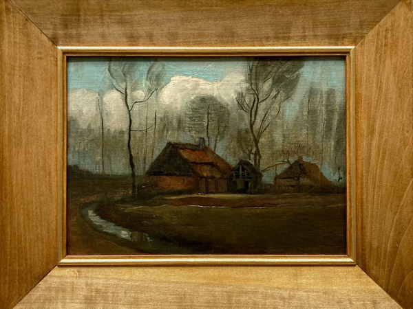 A painting in wide light brown wooden frames. It depicts two village cottages and several trees. Dark colors dominate